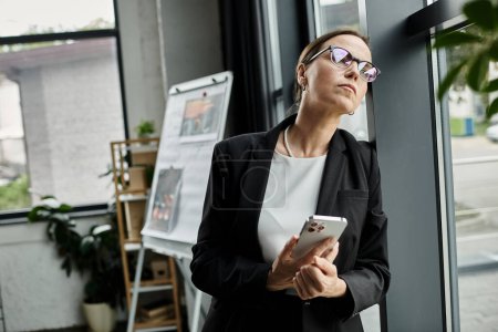 Téléchargez les photos : A middle-aged businesswoman looks worriedly at her phone by a window. - en image libre de droit