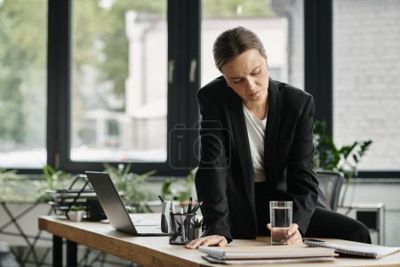 Middle-aged woman sits at desk, contemplating, holds glass of water.
