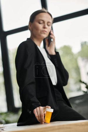 Middle-aged woman in office, multitasking with phone and pill bottle.