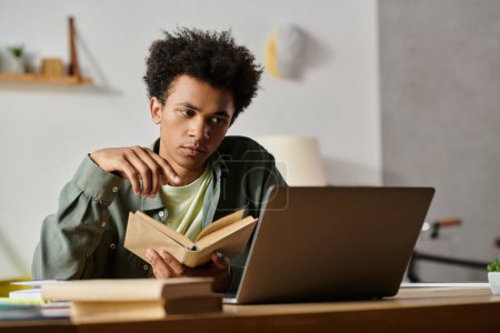 Young man engrossed in a book, studying at a table with a laptop.