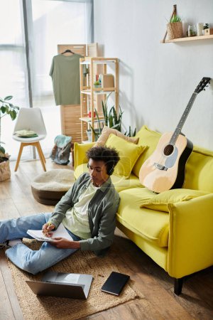 Young man, African American, sits near yellow couch with guitar.