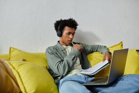 A young man, studying online with laptop and headphones, sits on a yellow couch.