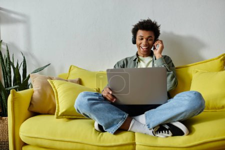 Young African American man studying online on a yellow couch with a laptop.