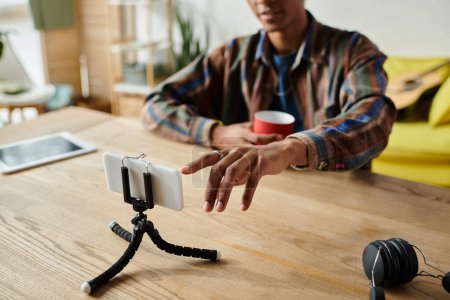 Young African American male blogger records himself on phone camera placed on a tripod at a table.