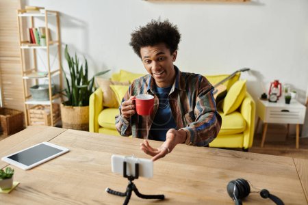 A young African American male blogger holding a cup of coffee while talking on a phone camera at a table in front of a yellow couch.
