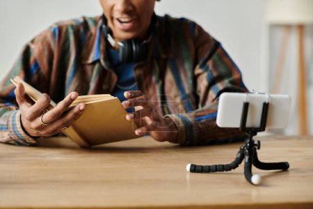 A young African American man reads a book while conversing on his phone camera at a table.