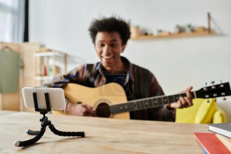 A young African American male blogger playing guitar on a tripod while speaking into a cell phone camera.