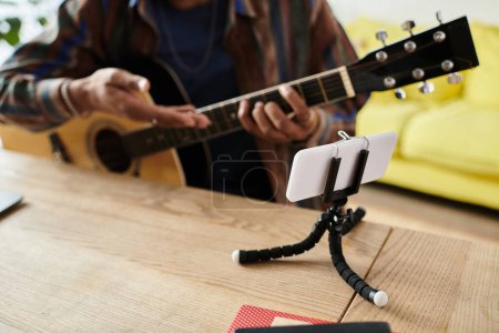 Foto de Young African American blogger playing an acoustic guitar on a tripod while talking on his phone camera. - Imagen libre de derechos