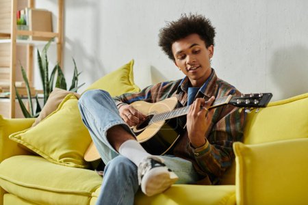 A young African American man playing an acoustic guitar on a yellow couch while talking on his phone.