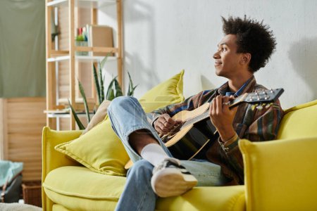 Foto de A young African American man plays an acoustic guitar while seated on a yellow couch. - Imagen libre de derechos