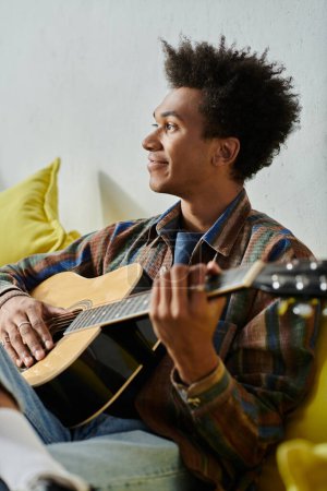 Young African American man strums acoustic guitar on couch.