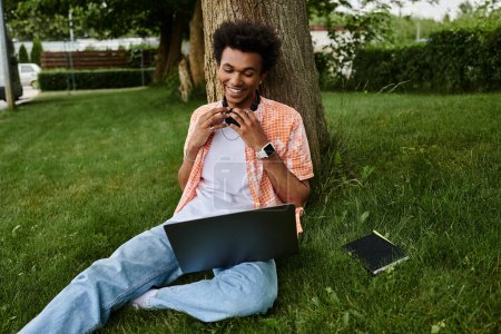 A young african american man seated on grass, engrossed in his laptop.
