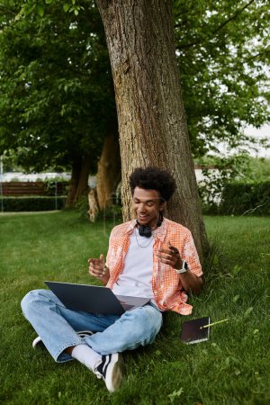 Young man, of African descent, using laptop while sitting under tree in park.