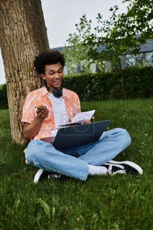 A young African American man sits under a tree, working on his laptop.