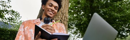 Téléchargez les photos : A young African American man holding a laptop in front of a tree in a park. - en image libre de droit