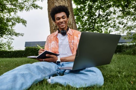 Téléchargez les photos : A young African American man sits on the grass, focused on his laptop. - en image libre de droit