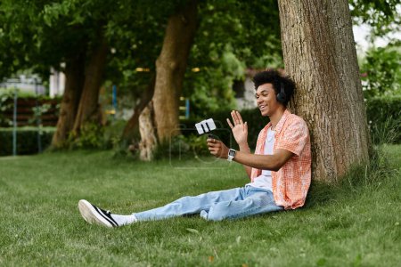 Foto de Young african american man sitting on grass, lost in music. - Imagen libre de derechos