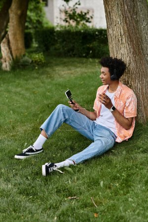A young African American man sitting under a tree, engrossed in his phone.