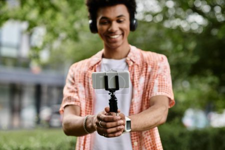 Photo for A young African American man holds a selfie stick in a park. - Royalty Free Image