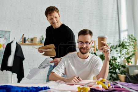 Ein schwules Paar arbeitet gemeinsam an einem Tisch, umgeben von Papieren und Tassen, während sie in ihrer Designer-Werkstatt trendige Kleidung kreieren.