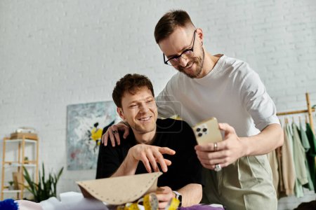 Foto de Dos hombres, una pareja gay amorosa, se paran orgullosamente uno al lado del otro en su taller de diseño, mostrando sus creaciones de atuendos de moda. - Imagen libre de derechos