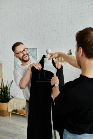 Foto de Dos hombres trabajan juntos en un taller de diseño, uno sosteniendo una camisa negra mientras que el otro la ajusta. - Imagen libre de derechos