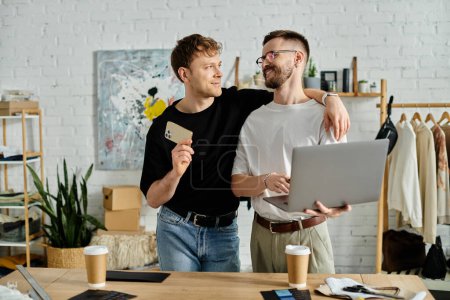 Foto de Dos hombres, una pareja gay, están en un taller de diseño, enfocados en la pantalla de un portátil mientras discuten la creación de atuendos de moda. - Imagen libre de derechos