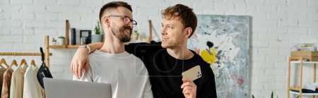 Foto de Dos hombres, una pareja gay, se paran uno al lado del otro en un taller de diseño, enfocado en crear atuendos de moda. - Imagen libre de derechos