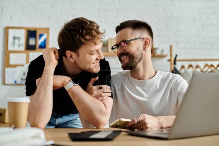 Foto de Dos hombres, una pareja gay, se sientan juntos en una mesa, enfocados en la pantalla de un portátil en un taller de diseño. - Imagen libre de derechos