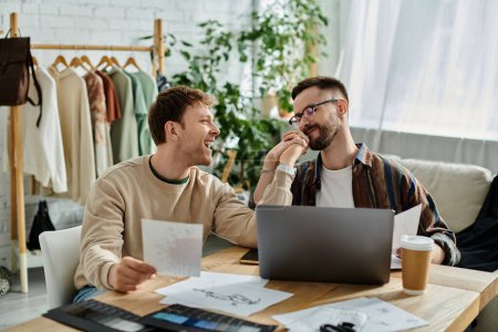 A gay couple collaborates on designing trendy attire at a table with a laptop.