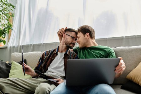 Téléchargez les photos : Deux hommes assis sur un canapé, se concentrant intensément sur un ordinateur portable pendant qu'ils travaillent sur la conception de vêtements à la mode ensemble. - en image libre de droit