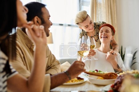 Foto de Un grupo diverso de amigos compartiendo una comida alrededor de una mesa, incluyendo una pareja lesbiana cariñosa. - Imagen libre de derechos