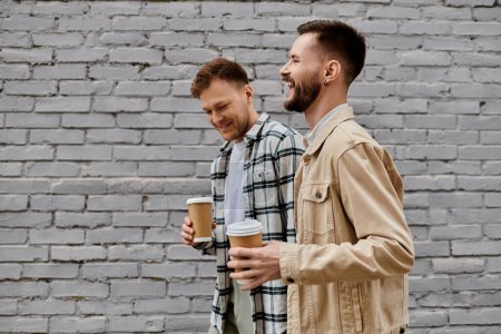 Foto de Una feliz pareja gay en traje acogedor disfrutando de café recién hecho al aire libre. - Imagen libre de derechos