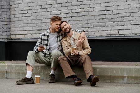 Two men in casual attire sitting happily together by a brick wall.