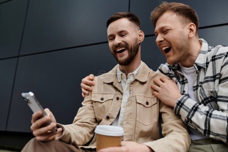 Zwei Männer in lässigen Outfits lachen zusammen, während sie auf ein Handy schauen.