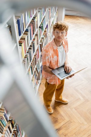 Photo for Young man stands by bookshelf, holding laptop. - Royalty Free Image