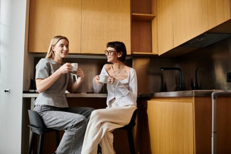 Foto de Una joven pareja de lesbianas disfruta del café en una acogedora cocina, charlando y compartiendo un momento en el mostrador. - Imagen libre de derechos