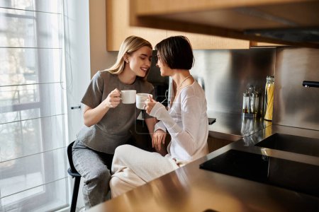 A young lesbian couple bonded over coffee cups, sitting close and chatting in a cozy kitchen inside a hotel room.