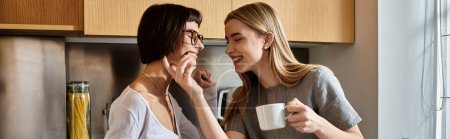 Una joven pareja lesbiana sosteniendo tazas de café, charlando y de pie en una elegante cocina de una habitación de hotel.