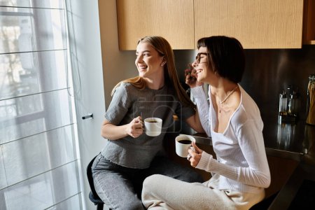 Young lesbian couple enjoy morning coffee together while sitting on window sill inside hotel room.