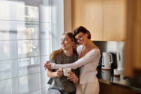 Foto de Dos mujeres jóvenes, una elegante pareja de lesbianas, se paran una al lado de la otra en una moderna cocina del hotel, sosteniendo tazas de café. - Imagen libre de derechos
