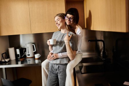 Foto de Dos mujeres, una joven pareja lesbiana, se paran una al lado de la otra en una acogedora cocina, cada una sosteniendo una taza de café. - Imagen libre de derechos