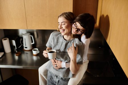 A young lesbian couple sitting together, sipping coffee in a cozy hotel room kitchen.
