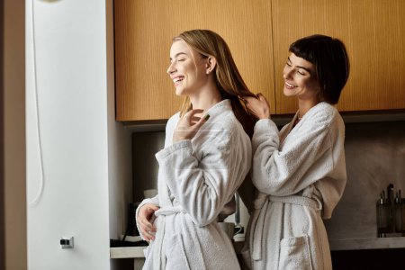 Téléchargez les photos : Un jeune couple de lesbiennes debout dans une cuisine confortable portant des peignoirs de bain. - en image libre de droit