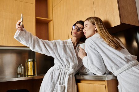 Two young women, dressed in bath robes, happily take a selfie together in a modern kitchen setting.