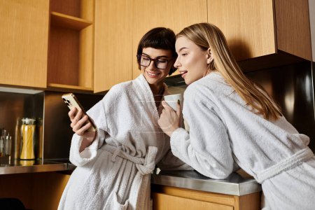 A young lesbian couple in bath robes share a laugh while standing next to each other in a cozy kitchen.