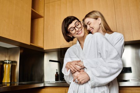 Un jeune couple lesbien en peignoir partage un moment de joie tout en se tenant ensemble dans une cuisine chaleureuse et accueillante.