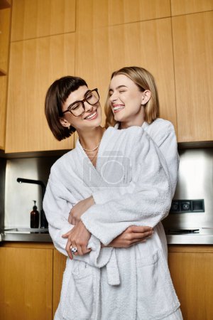 Téléchargez les photos : Un jeune couple lesbien en peignoir se tient à proximité dans une cuisine confortable, partageant un moment privé et tendre. - en image libre de droit