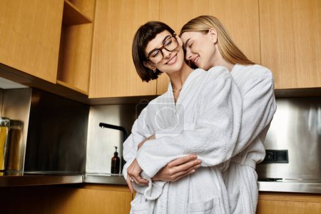 Photo for Two young women in bath robes, standing side by side, smiling in a contemporary kitchen. - Royalty Free Image