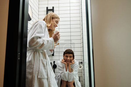 A woman in a bathrobe brushes her teeth while her partner watches in a hotel bathroom.
